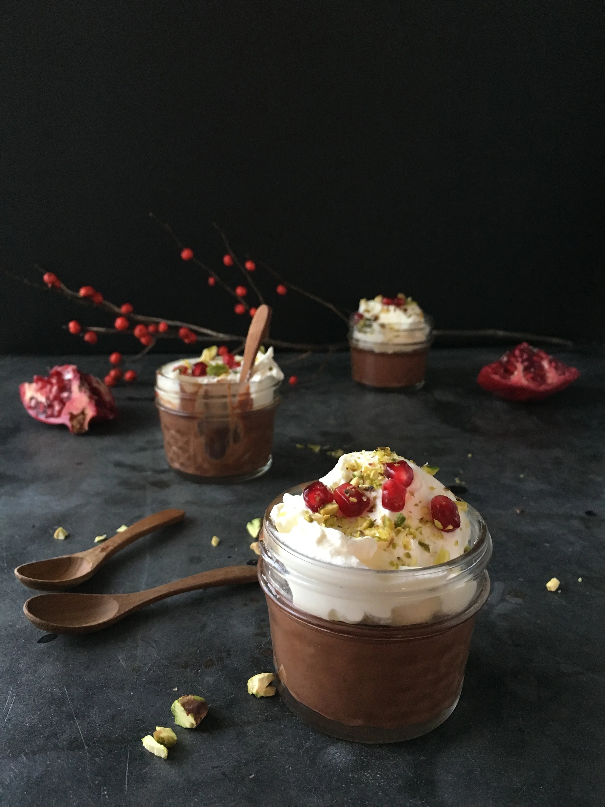 Two small glass jars of chocolate mousse topped with whipped cream, pomegranate seeds and crushed pistachios on a dark moody tabletop