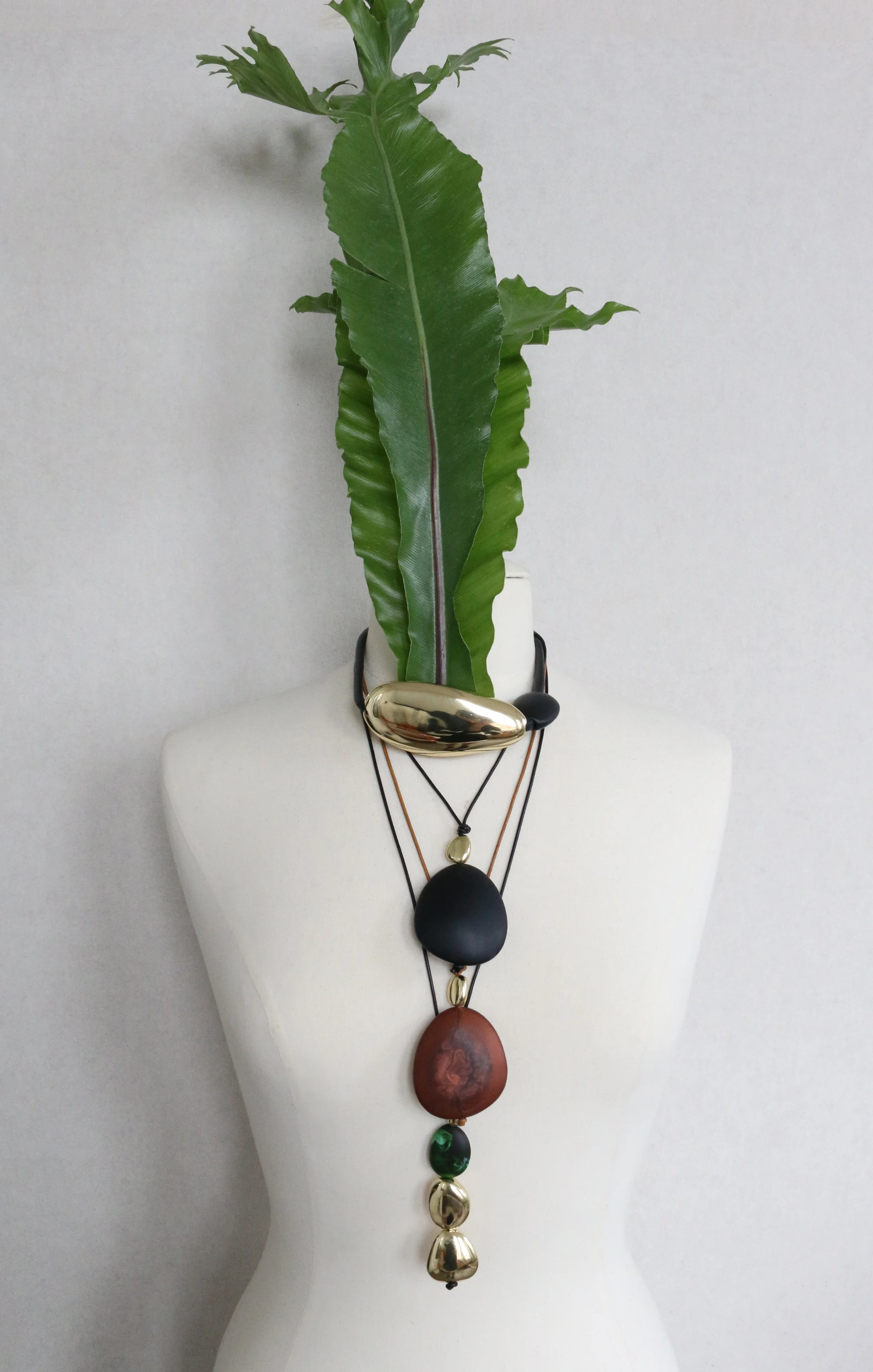 Mannequin torso wearing a layered necklace of black, gold and reddish-brown beads accented with large green tropical leaves against a plain light backdrop